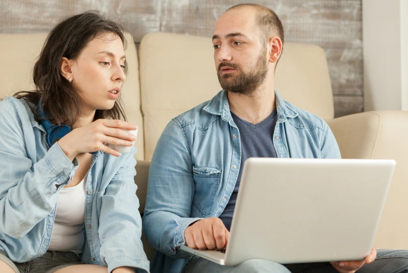 Woman holding a coffee cup talking to a bearded man with a laptop on a couch, a couple in a serious discussion about deciding on a vasectomy.