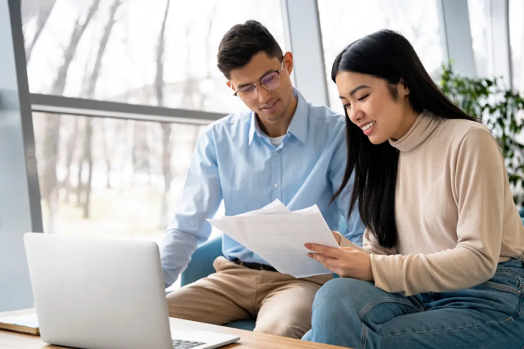 Man in glasses and woman in a beige top reviewing printed documents together beside a laptop comparing vasectomy clinic costs.