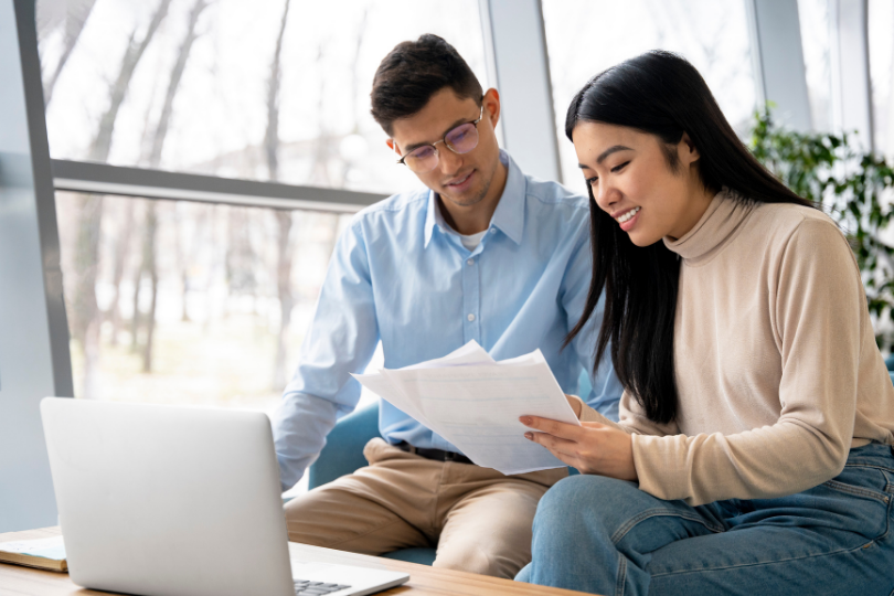 Man in glasses and woman in a beige top reviewing printed documents together beside a laptop comparing vasectomy clinic costs.