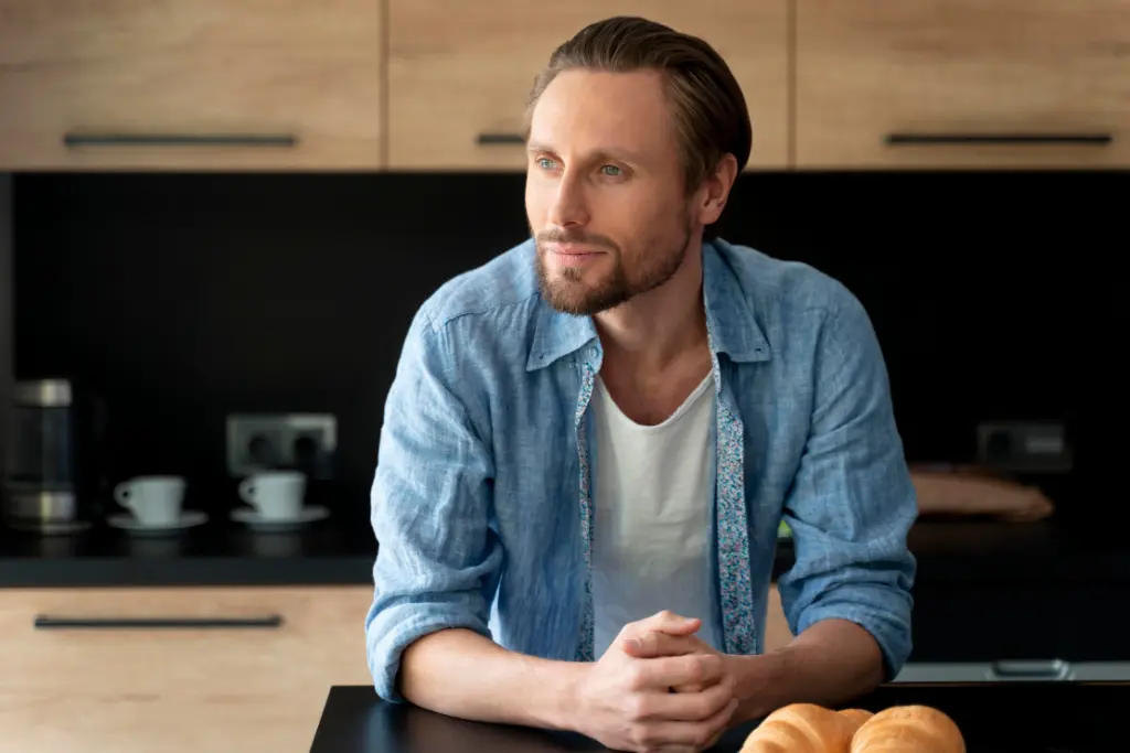 Single man with blue eyes and a beard standing at a kitchen counter — considering sperm freezing as a personal decision.