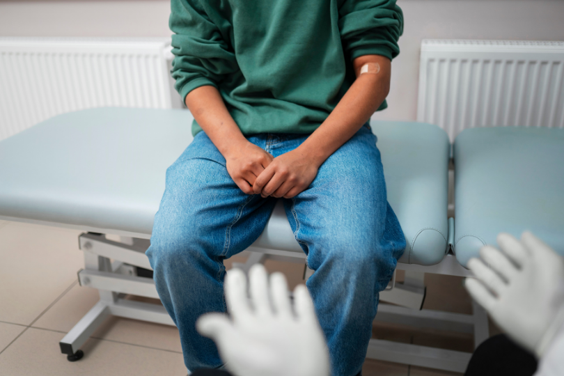 Male patient in jeans and a green sweater with an arm bandage sitting on an exam table while a gloved doctor faces him, a vasectomy consultation in progress.